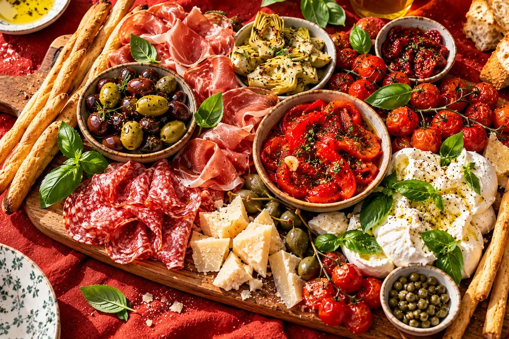 A rustic board with folded salami, marinated peppers, olives, mozzarella, grissini and tomatoes
